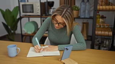 Ofis masasındaki bir defterde yazan bir kadın. Yanında cep telefonu olan, iş yerinde çalışan sarışın bir kadın..
