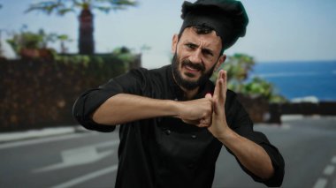 Siyah üniformalı İspanyol şef, güneşli bir sokakta yumruklarıyla enerji dolu jestler yapıyor. Palmiye ağaçları ve mavi gökyüzü arkaplanı arasında kararlılık ve duyguyu gösteriyor..