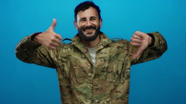 Ordu üniformalı İspanyol bir adam, sakallı, mavi arka planda bir aşağı bir yukarı hareket ediyor, modern asker hayatında karışık duygular ve ikiliği ifade ediyor..