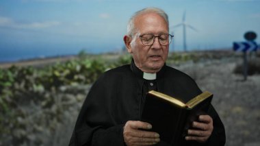 Arka planda yel değirmenleri olan bir açık alanda kitap tutan kıdemli bir rahip, dinsel bir açık hava manzarasını ve huzurlu bir kırsal ortamı gösteriyor..