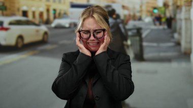 Arka planda bulanık yayalar ve şehir araçlarıyla çevrili, gözlüklü ve koyu renk ceketli, hareketli bir caddede duygusal olarak ağlayan bir kadın..