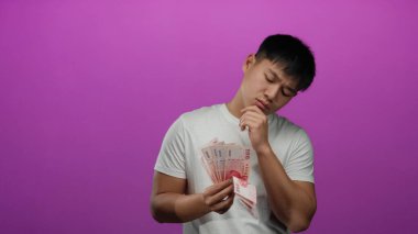 Tayvan dolarlarını ihtiyatlı bir şekilde pembe arka plan duvarının üzerinde tutan genç adam derin düşüncelere dalmış bir ifadeyle ve sıradan bir pozla Asya para birimini gözler önüne seriyor.
