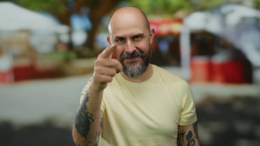 Güneşli bir günde sokak ortamında gülümseyen bir adam sarı bir gömlek giyerken sakalını, kelliğini ve dövmelerini sergiliyor..