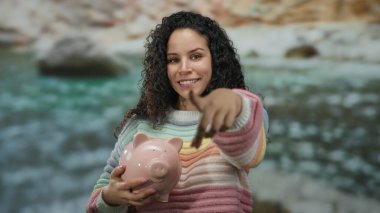 Sahilde elinde pembe bir kumbarayla duran, arka planda okyanus bulunan renkli süveter giyen bir kadın, dışarıda mali güvenini ve mutluluğunu gösteren kameraya bakıyor..