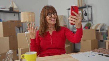 Gülümseyen ve selfie çeken bir kadın, modern bir iç mekanı olan parlak, yeni bir evde kutuları taşıyor..
