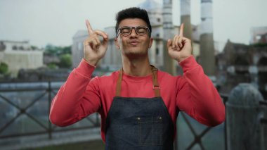 Gözlüklü ve kırmızı tişörtlü genç adam şehir caddesinde ellerini kaldırmış, bulanık mimari geçmişi ve kendine güveni ve neşesini sergiliyor..