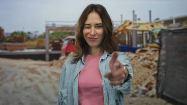 İspanyol kadın inşaat alanında gülümsüyor, günlük kıyafetler giyiyor, bulanık bina makineleri ve açık hava düzeneği ile canlı, genç bir atmosferi taşıyor..