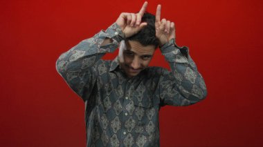 Şablonlu gömlekli genç adam elleri kırmızı arka plana karşı boynuz gibi şakacı hareketler yapıyor. Yaratıcılık ve canlı, izole bir ortamda günlük İspanyol tarzı sergiliyor.