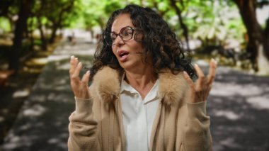 Kıvırcık saçlı ve gözlüklü olgun bir kadın, verimli yeşilliklerle çevrili güneşli bir parkta huzurlu bir anın tadını çıkarıyor..