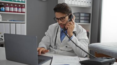 Tıbbi malzemeler ve organize dosyalarla çevrili bir dizüstü bilgisayar üzerinde çalışırken klinik odasında danışmanlık yapan genç İspanyol erkek doktor profesyonel bir ayar sergiliyor..