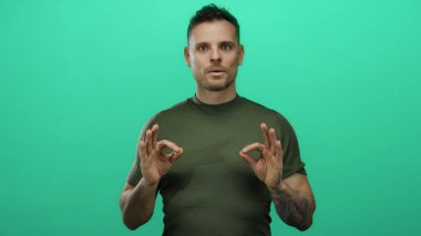 Genç İspanyol adam, izole edilmiş yeşil arka plana karşı meditasyon yapıyor. Sakince ifadesini ve dövmelerini gösteriyor. Rahatlama ve farkındalık taşıyor.