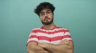 Mavi arka planda kollarını kavuşturan gözlüklü ve sakallı İspanyol genç adam çizgili gömlek giyiyor sakin bir ifade sergiliyor..