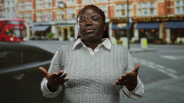 Arka planda ikonik kırmızı bir otobüsle şehir caddesinde duran gözlüklü ve gri kazaklı bir kadın..