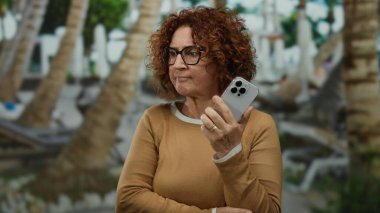 Havuz başında akıllı telefonları olan kıvırcık saçlı günlük giysiler giyen bir kadın açık hava otel ortamında modern teknoloji kullanımını yansıtıyor..