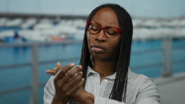 Gözlüklü bir kadın, arka planda teknelerle sahil limanında bilek ağrısı çekiyor açık hava manzarası sunuyor ve sağlık ve rahatlamaya odaklanıyor..