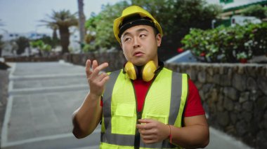 Sarı kasklı ve yelekli bir adam şehir sokağında bitkilerle dikiliyor, güneşli bir günde inşaat kıyafetleri ve aksesuarları sergiliyor..