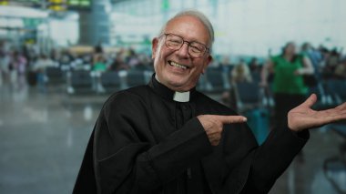 Siyah cüppeli kıdemli rahip gülümsüyor ve bulanık gezginler ve oturanlarla dolu bir havaalanı arkaplanını işaret ediyor..