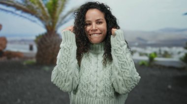 Parkta gülümseyen bir kadın palmiye ağaçlarının altında rahat bir süveter giyiyor, gündüz doğal ortamında neşeli bir ifade sergiliyor..