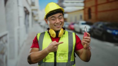 Güler yüzlü Asyalı adam emniyet yeleği giyiyor ve kentsel caddedeki elektronik cihaza nokta koyuyor. Arka planda arabaları var. Şehir temalı açık hava portresi çizimleri yapıyor.