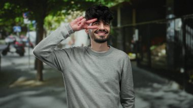 Genç adam gülümsüyor ve açık havada ağaçların gölgesinin altında bir barış işareti yapıyor, gri bir kazak giymiş, rahat ve kendinden emin bir tavır sergiliyor..