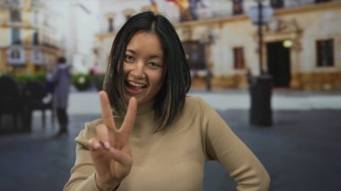 Kadın gülümsüyor ve açık hava sokak sahnesinde barış işareti ile poz veriyor, gençlik enerjisini ve günlük kıyafetlerle şehir tarzını gösteriyor..