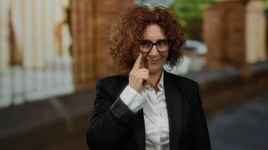 Woman gesturing confidently wearing glasses on city street with blurred urban background showing diverse middle-aged style outdoors.