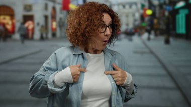 İspanyol orta yaşlı, kıvırcık saçlı ve gözlüklü bir kadın şehir caddesinde şaşırmış, günlük kıyafetler giymiş, kendini işaret ediyor..