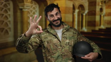 Askeri üniformalı ve sakallı bir adam katolik kilise ortamında, dekoratif mimari ile çevrili bir ortamda, güven duygusunu dile getiriyor..