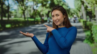 Mavi kazaklı genç kadın güneşli bir parkta, ağaçlarla çevrili ve bulanık insanları işaret ediyor..