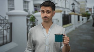İspanyol bir adam bir şehir caddesinde kahve fincanı tutarken senin jestini izliyor, mizahi ve sıradan bir şehir manzarası ve kahve kültürü aktarıyor..