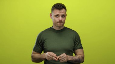 Yeşil gömlekli genç İspanyol adam izole edilmiş sarı arka planda dalgın görünüyor parlak ışık altında çekici özellikler gösteriyor ve kolunda dövmesi var..