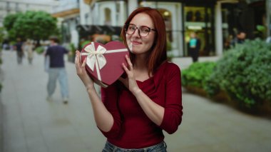 Şehir caddesinde elinde kalp şeklinde bir hediye kutusu olan bir kadın gözlüklü ve kırmızı tişörtlü. Etrafı yeşilliklerle çevrili ve arka planda yürüyen insanlar.