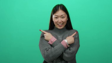 Kadın gülümserken, canlı yeşil arka plana karşı işaret ederken rahat bir süveter ve pembe saat giyerek neşeli ifadeyi ve modern tarzı ön plana çıkarıyor.