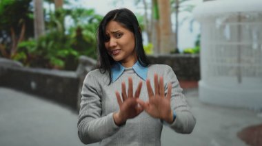 Sokakta yeşillik içinde bir kadın, dur hareketi yapıyor, kaygısını dile getiriyor, günlük süveter giyiyor, gün boyunca şehir merkezini ön plana çıkarıyor..