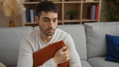 Evdeki bir kanepede oturan genç adam, düşünceli görünüyor ve yastık tutuyor, etrafı raflar ve dekorlarla çevrili, rahat bir oturma odasında..