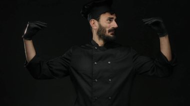Sakallı İspanyol erkek aşçı siyah eldiven giyip siyah bir arka plana karşı üniforma giyiyor. Mutfak uzmanlığı ve tutkusunu aktarıyor..