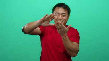 Kırmızı gömlekli genç adam yeşil arka plana karşı para hareketi yapıyor. İzole edilmiş bir stüdyoda başarıyı ve finansal refahı sembolize eden neşeli bir ifadeyle.