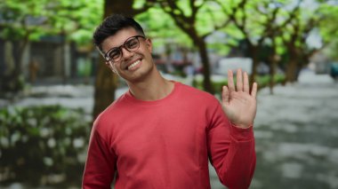 Gözlüklü genç adam gülümsüyor ve gün içinde ağaçlarla dolu canlı bir şehir sokağında el sallıyor..