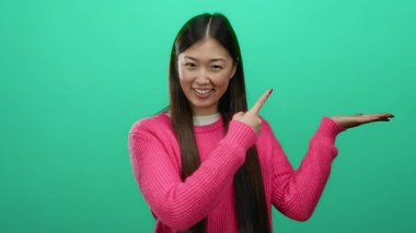 Pembe kazaklı kadın, parlak yeşil bir arka planda el kol hareketi yapıyor, etkileyici bir poz sergiliyor ve çekici bir gülümseme sergiliyor, neşeli bir ortamda kendine güveni ve coşkuyu somutlaştırıyor..