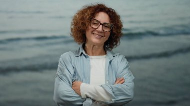 Orta yaşlı, esmer, kıvırcık saçlı ve gözlüklü İspanyol kadın kollarını çapraz bir zemine dayamış gülümsüyor, kendine güveni ve sıcaklığıyla..