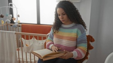 Renkli kazaklı kitap okuyan bir kadın, evde beşiğin yanında oturur, ebeveynlik ve rahatlık odaklı samimi ev ortamlarını yakalar..