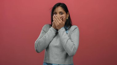 Gri kazaklı, uzun koyu renk saçlı bir kadın kırmızı duvara karşı ağzını kapatıyor. Şok duygusunu ve izole atmosferi gösteriyor..