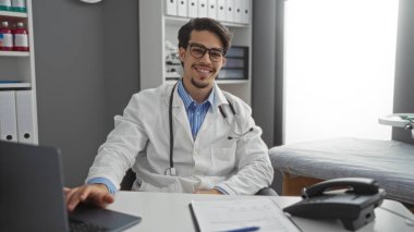 Genç İspanyol erkek doktor hastane odasında tıbbi ekipman ve belgelerle güvenle gülümsüyor. Profesyonellik ve karşılayıcı bir atmosfer sergiliyor..
