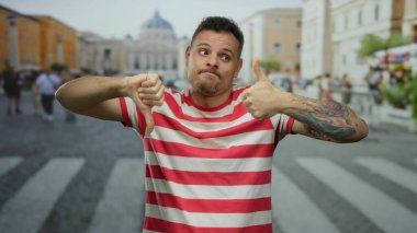 Çizgili gömlekli genç adam şehir sokaklarında karışık başparmak hareketleri ve şehir ortamında dövmeli kol gösteriyor..