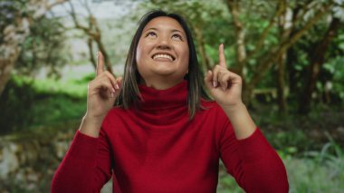 Açık bir gökyüzünün altında ağaçlarla kaplı yeşil bir parkı işaret eden kırmızı kazaklı bir kadın açık hava ortamında iyimserlik ve neşe öneriyor..