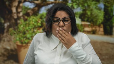 Parkta ağzı, gözlüğü ve beyaz gömleği olan, yeşilliklerle çevrili, açık hava yaz ortamında duyguları somutlaştıran bir kadın iğreniyor..