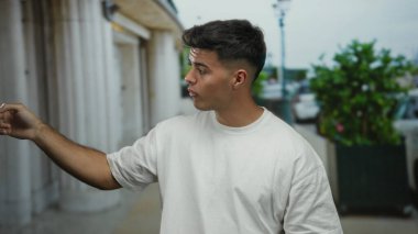 Beyaz gömlekli genç adam hareketli şehir caddesinde köşe yazıları ve yeşilliklerle dolu, şehir ortamında ifade ve etkileşim sergiliyor..