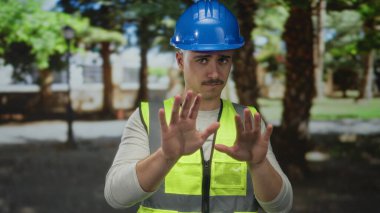 Yansıtıcı yelek ve şapka giyen genç İspanyol bir adam açık hava parkında reddedilme jesti yapıyor..