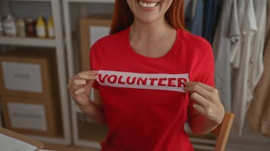 Bağış merkezinde kırmızı gönüllü gömleği sergileyen bir kadın. Arka planda malzeme rafları var. Kamu hizmeti ve destek ruhunu vurguluyor..