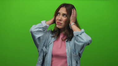 Canlı yeşil bir arkaplan önünde, kulaklarını sıkıntılı bir ifadeyle kapatan kadın, sıradan bir kot ceket giyiyor, stres ve hüsranın sinyallerini veriyor..